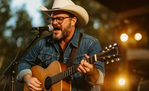 Man in cowboy hat and denim jacket singing and playing guitar outdoors.