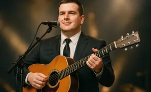 A man in a suit plays an acoustic guitar and sings into a microphone on stage.