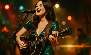 Singer Kacey Musgraves performs live, playing her guitar and singing into a microphone, illuminated by stage lights.