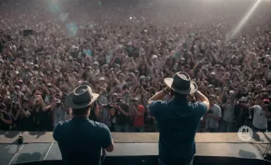 Two men in hats on a stage face a massive, cheering crowd at an outdoor concert.