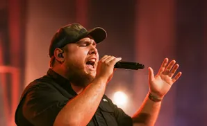 A man in a camo hat sings into a microphone with his hand raised.