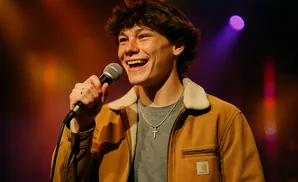 A young man singing into a microphone on stage under warm stage lights.
