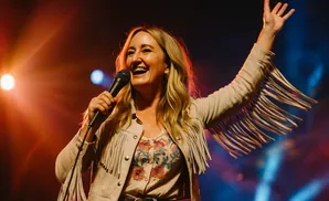 A woman in a fringed jacket sings into a microphone on a stage with colorful lighting.