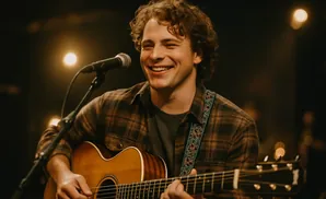 A man with curly hair smiles while playing an acoustic guitar and singing into a microphone.