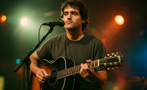 A man with dark curly hair plays an acoustic guitar and sings into a microphone on a dimly lit stage.