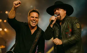 Two men rock out on stage, one singing with a microphone, the other playing a guitar and raising a fist.