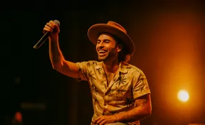 A man in a hat and floral shirt sings into a microphone on a stage with warm lighting.