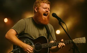 A red-bearded man sings into a microphone while playing a resonator guitar.
