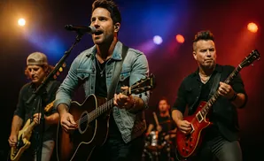 A male country singer plays an acoustic guitar and sings into a microphone on stage, with bandmates playing guitar and drums behind him.