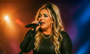 A woman with blonde hair, wearing a black top and gold necklace, sings into a microphone on stage.