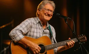 A smiling man with gray hair and glasses plays an acoustic guitar into a microphone.