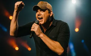 A man in a cap sings into a microphone with his fist raised, bathed in stage lights.