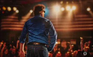 Man in a blue shirt and jeans facing away from camera on a stage, American flags and audience in background.
