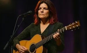 A woman with red-streaked hair plays an acoustic guitar on stage, singing into a microphone.