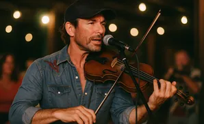 Man wearing a baseball cap and denim shirt plays the violin into a microphone, with blurred lights in the background.