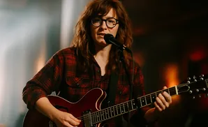 A woman with glasses and red plaid shirt plays an electric guitar while singing into a microphone.