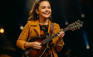 A woman with brown hair in a ponytail plays a mandolin onstage, wearing a yellow jacket and floral dress.