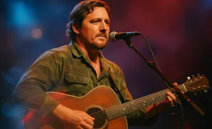 A man plays an acoustic guitar and sings into a microphone on a stage with blue and red lighting.