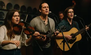 A woman plays the violin while two men play guitar and mandolin, singing into microphones.