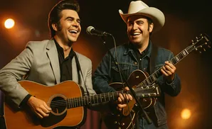 Two men play guitars and sing into microphones on a dimly lit stage.