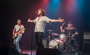 A band performs on stage with a singer in the center, a guitarist on the left, and a drummer on the right.