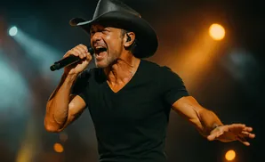 Country singer Tim McGraw in a black cowboy hat and t-shirt sings into a microphone on stage.