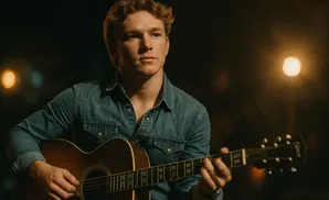 A man with wavy brown hair plays an acoustic guitar in dim lighting, wearing a denim shirt.