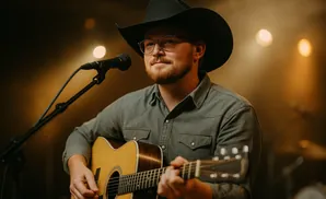 Man in a cowboy hat and glasses plays an acoustic guitar on stage, with spotlights in the background.
