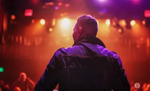 A man in a denim jacket stands with his back to the camera on a stage, bathed in red and purple stage lights.