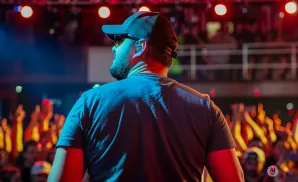 Man in a baseball cap and sunglasses on stage, facing away from the camera, with a cheering crowd in the background.