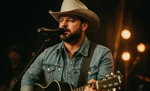 A man in a cowboy hat and denim jacket plays an acoustic guitar and sings into a microphone.