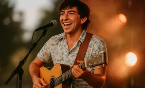 A smiling man plays an acoustic guitar and sings into a microphone, bathed in warm stage lighting.