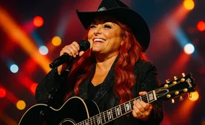 Wynonna Judd smiles while singing into a microphone and playing a guitar on stage.