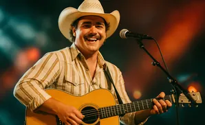 Man wearing a cowboy hat and a striped shirt plays an acoustic guitar and sings into a microphone.
