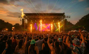 A crowd cheers at a nighttime outdoor music festival with a stage, lasers, and pyrotechnics.