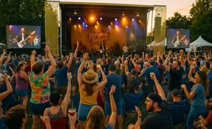 People with raised hands watch a band perform on a stage at an outdoor festival.