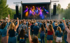 People at an outdoor concert, facing a stage lit with purple and orange lights. Many hands are raised in the air.