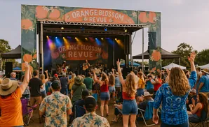 People enjoy an outdoor concert, raising their hands in enthusiasm before a stage decorated with oranges.