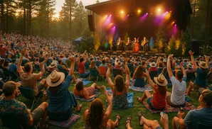 Outdoor concert with band on stage, audience sits on grass in front of a stage at sunset.