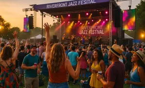 People with raised hands enjoy a band performing on stage at the Riverfront Jazz Festival at sunset.