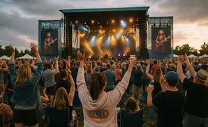 Crowd with hands raised at an outdoor concert, with a stage and screens showing performers.