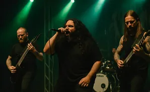 A metal band performs on stage, with a vocalist singing into a microphone and two guitarists playing.