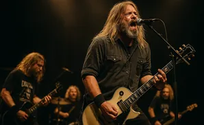 A lead singer and guitarist with long blonde hair and a beard, singing into a microphone and playing a white electric guitar.