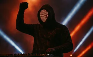 A person in a hooded sweatshirt and a mask with glowing red lights operates DJ equipment, with dramatic red and blue stage lights behind them.