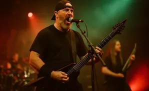 A man with a beard and a baseball cap sings into a microphone while playing a black electric guitar on stage.