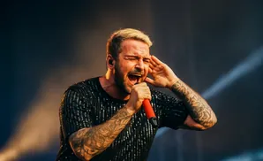 Man with blonde hair and tattoos sings passionately into a red microphone, holding his other hand to his ear.