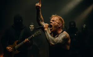 A male singer with blonde hair and tattoos screams into a microphone while other masked figures play instruments behind him.