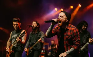 A male singer with a red beard and plaid shirt sings into a microphone on a stage with bandmates.