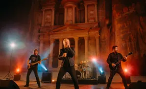 A band performs a concert in front of a large ancient stone facade, illuminated by stage lights and fog.