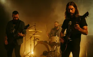A band performs on stage with bright lights and fog. The guitarist in the foreground plays an electric guitar, while a bassist and drummer are visible i…